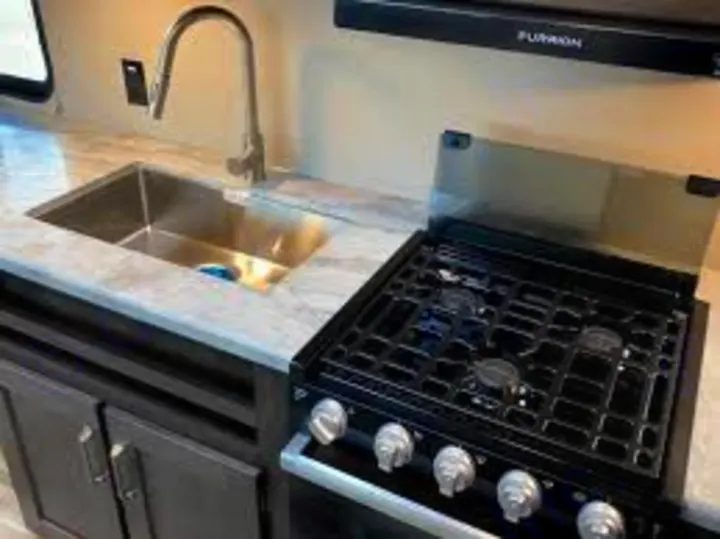 RV rental kitchen: Features a stainless steel sink with faucet on light counter, plus a black four-burner stove/oven combo on the right.. Grand Design Transcend Xplor 2021