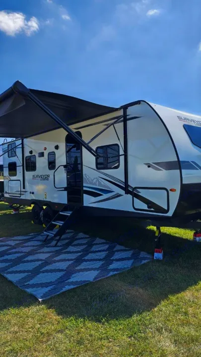 This RV rental is set up for camping, with an awning and mat on green grass under a blue sky. Perfect for outdoor travel.. Forest River Surveyor Legend 2024