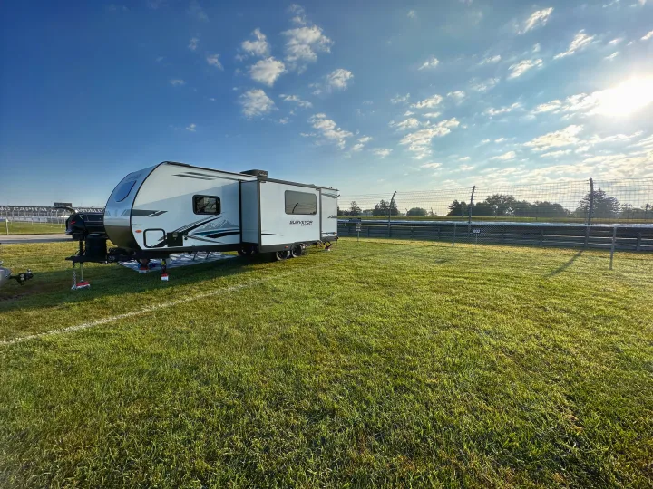 An RV rental trailer is parked on a dewy grass field next to a race track on a sunny morning.. Forest River Surveyor Legend 2024