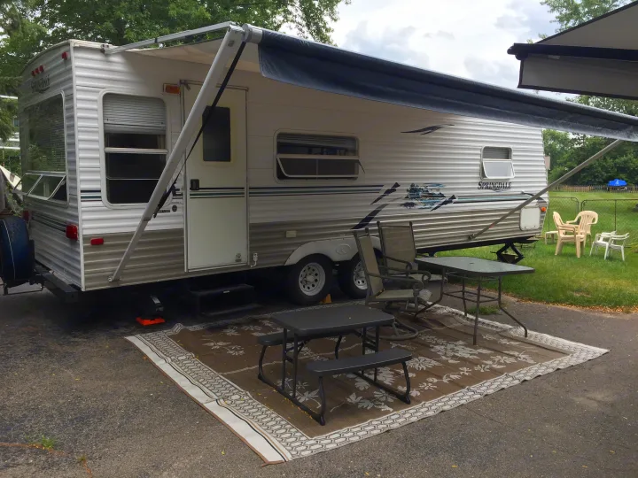 This RV rental, a Springdale travel trailer, is set up at a campsite with an extended awning, outdoor table, benches, and chairs on a patio rug for comfortable outdoor living.. Keystone RV Springdale 2005