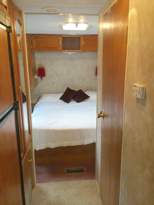 View into an RV rental bedroom: white bed with dark pillows, wall lamps, and wood cabinetry, seen through an open doorway.. Keystone RV Springdale 2005