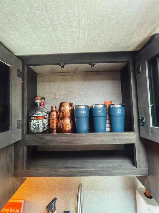 RV rental kitchen cabinet showing organized drinks: Espolon tequila, copper mugs, and blue Yeti tumblers.. Grand Design Transcend Xplor 2023