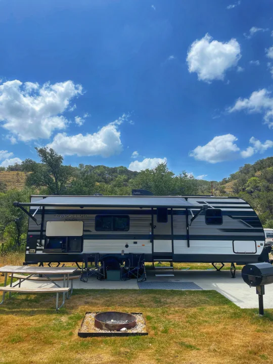 RV rental setup at a sunny campsite, featuring an extended awning, picnic table, and fire pit under a blue sky with clouds.. Grand Design Transcend Xplor 2023
