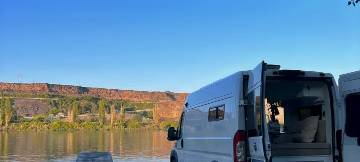 This RV rental offers stunning waterside views. Open doors reveal a cozy camper van interior by cliffs & clear skies. Ideal for travel.. Dodge Ram 1500 2019