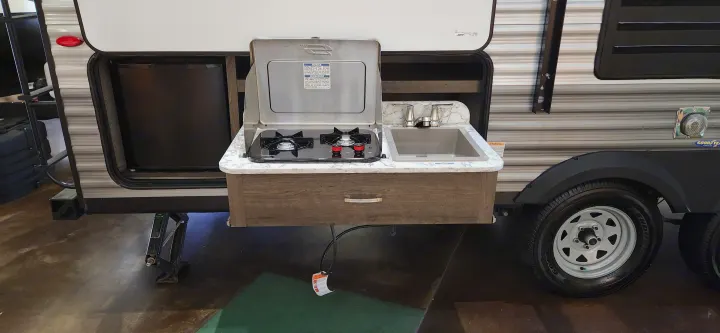 An RV rental's outdoor kitchen: a pull-out unit with a two-burner stove and sink, next to an accessible refrigerator.. Jayco Jay Flight SLX 2021