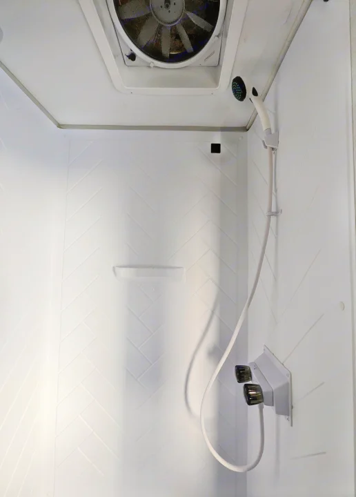 Compact white shower stall in an RV rental, showing a ceiling fan, handheld shower, and herringbone wall pattern.. Forest River Wildwood FSX 2021