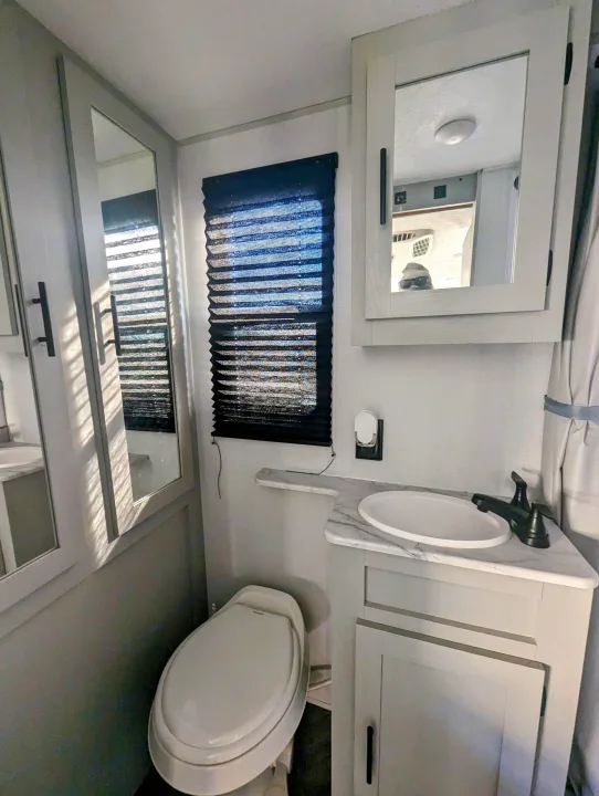 RV rental bathroom: toilet, sink, mirror reflecting an interior with a character in sunglasses, and window with blind.. Forest River Wildwood FSX 2021