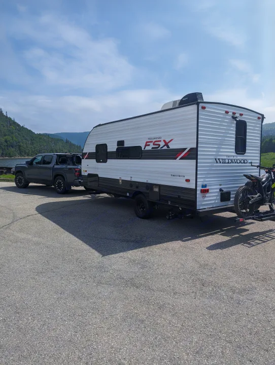 This RV rental, a Wildwood FSX travel trailer, is hitched to a truck, parked by a lake with mountains. A dirt bike is on the rear rack.. Forest River Wildwood FSX 2021