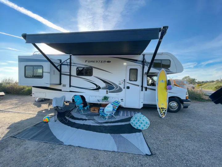 Sunny rv rental campsite features a white RV, extended awning, surfboard, chairs, and beach ball on a mat. Adventure awaits!. Forest River Forester 2020