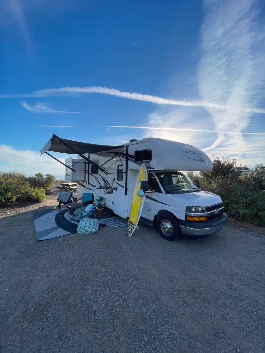 Scenic rv rental setup with a surfboard, chairs, and awning under a clear sky, near the ocean.. Forest River Forester 2020