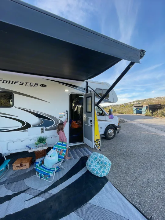 RV rental setup: A girl by a white RV with an extended awning, outdoor chairs, picnic basket, and a surfboard. Perfect for family camping.. Forest River Forester 2020