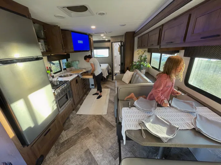 Inside a bright rv rental, a woman preps in the kitchen while a child sits at the dining table, likely preparing for a meal.. Forest River Forester 2020