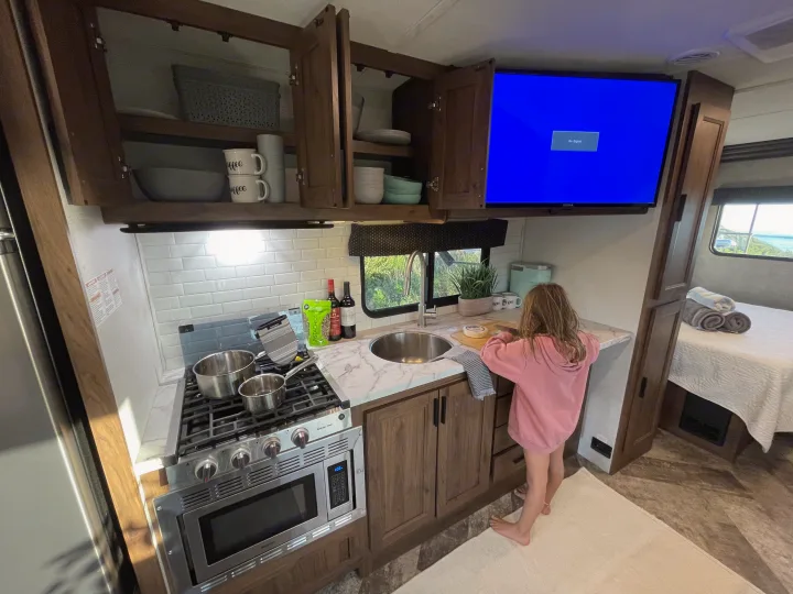 Child preps food in a compact rv rental kitchen, complete with stove, sink, open cabinets, and TV. Ideal camper setup!. Forest River Forester 2020