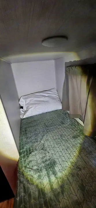 This RV rental features a compact sleeping bunk with a green quilted bedspread, white pillow, and overhead light.. Gulf Stream Ameri-Lite LE 2023