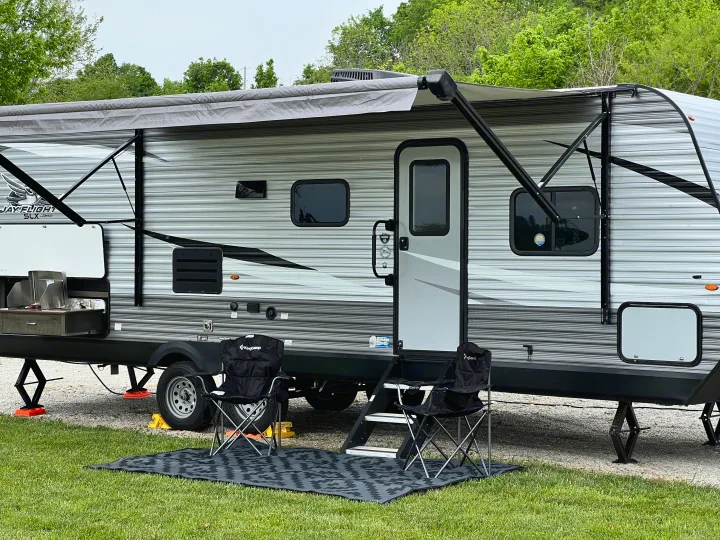 This RV rental is set up at a campsite with an extended awning, outdoor kitchen, chairs, and mat, ready for outdoor living.. Jayco Jay Flight SLX 2021