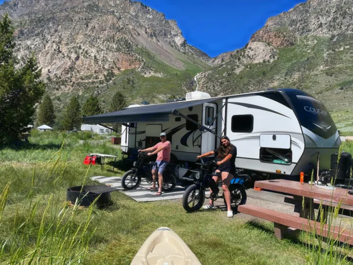 RV rental mountain camp: Two people and e-bikes at their travel trailer in a scenic campground.. Keystone RV Cougar 2021