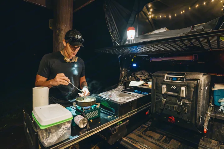 Nighttime RV rental setup: Man cooking outdoors with portable kitchen, fridge, and rooftop tent.. Jeep Gladiator 2022