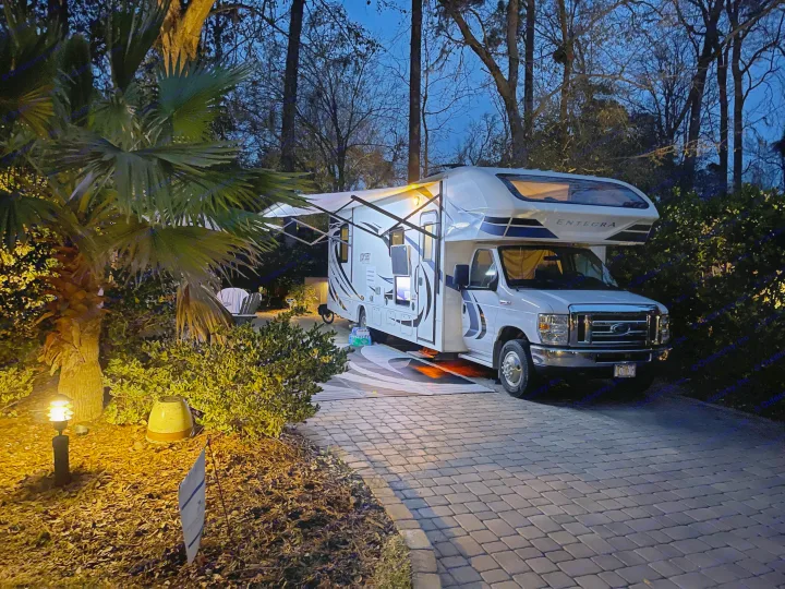 Evening view: A white Entegra RV rental with awning extended, parked on a brick driveway, illuminated by warm lights.. Entegra Coach Odyssey 2020