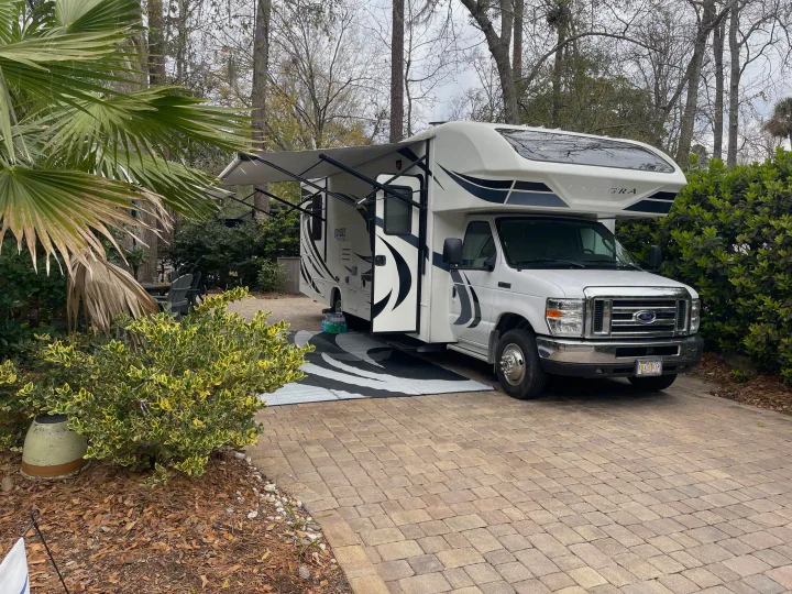 White RV rental parked on a paved site with extended awning, lush plants, and trees – ready for a road trip.. Entegra Coach Odyssey 2020