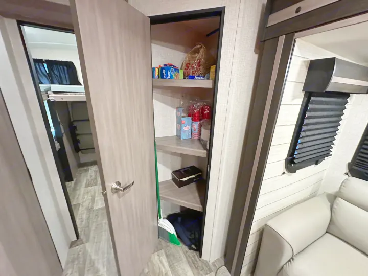 RV rental interior shows an open pantry stocked with essentials, a broom, and a glimpse of bunk beds and a sofa.. Crossroads RV Sunset Trail 2022