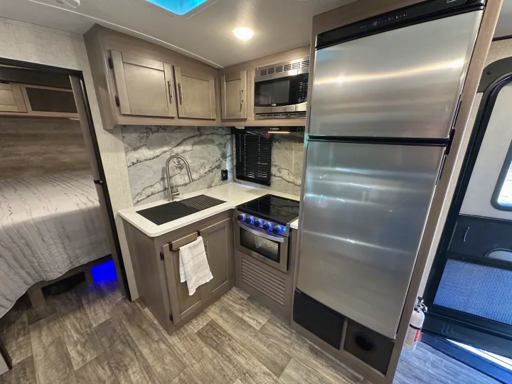 Inside this rv rental: A modern kitchen with stainless steel appliances, marble-look backsplash, and wood-look floors, beside a bed.. Crossroads RV Sunset Trail 2022