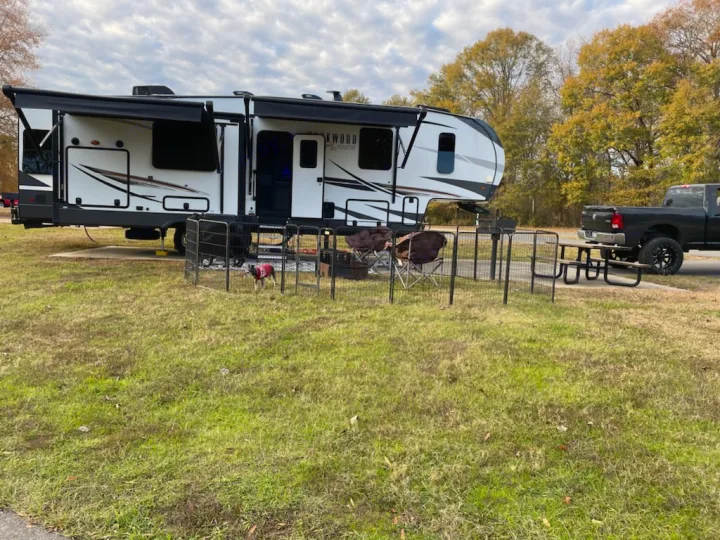 An RV rental is set up at a grassy campsite, featuring a dog pen with a small dog, chairs, picnic table, and a truck.. Forest River Rockwood Ultra Lite 2022
