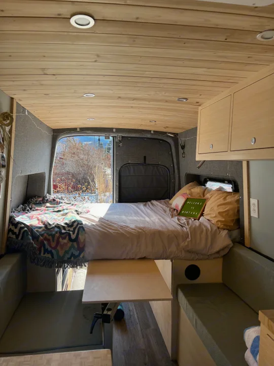 Cozy RV rental interior with a comfy bed, wood ceiling, seating, and "Welcome to Montana" sign.. Ford Transit 2022