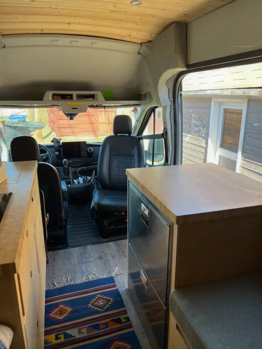 Spacious RV rental interior featuring warm wood cabinetry, built-in fridge, and a clear view to the driver's cockpit.. Ford Transit 2022