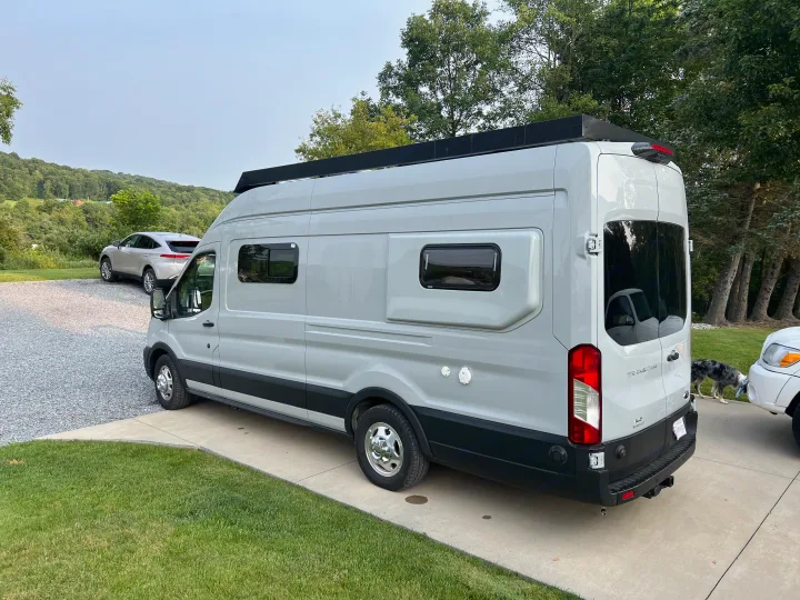 A light gray Ford Transit camper van, ready as an RV rental, is parked on a concrete driveway with hills & trees.. Ford Transit 2022