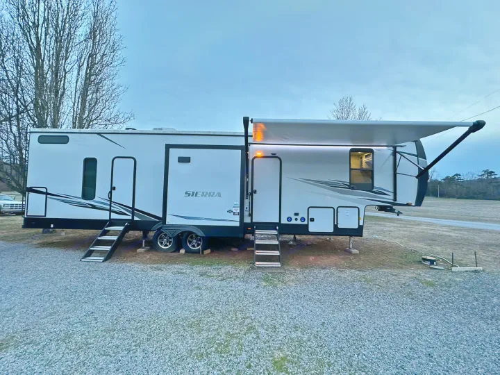 This RV rental is parked on gravel, with its awning extended and lights on. Bare trees frame the overcast sky.. Forest River Sierra 2022