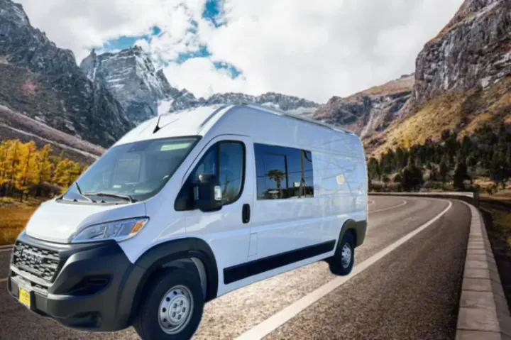 White RV rental van on a scenic mountain road with snow-capped peaks and autumn trees. Plan your adventure.. RAM PROMASTER 2500 2023