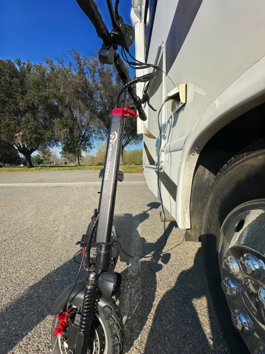 A black electric scooter charges from a white RV rental on a paved lot under a blue sky, ideal for campsite exploration.. Thor Four Winds 2019