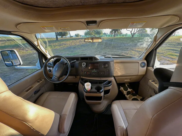 Interior view of an **RV rental's** front cabin, showing the dashboard and seats, looking out to a scenic park.. Thor Four Winds 2019