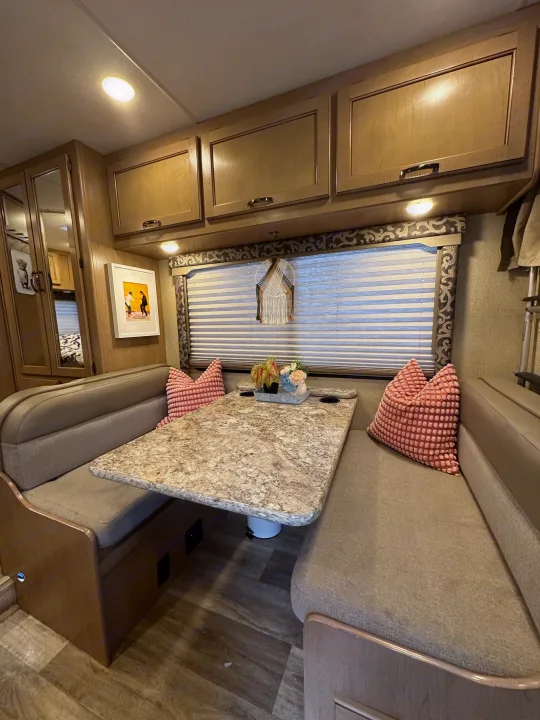 Cozy RV rental dinette setup with a stone-look table, patterned window blinds, and overhead wood cabinets.. Thor Four Winds 2019