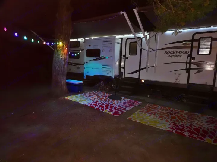 Illuminated RV rental at night with bright awning lights, colorful string lights on a tree, and patterned ground mats.. Forest River Rockwood Signature Ultra Lite 2015