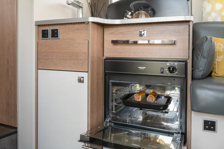 This RV rental kitchen features a compact oven baking sausage rolls, a fridge, sink, and UK outlets, ideal for travel.. GlamperRV DBL 2026