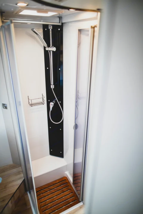 Detailed view of a compact RV rental shower cabin with sleek black fixtures, a wall-mounted shower, and a comfortable wooden mat.. GlamperRV DBM Automatic 2025
