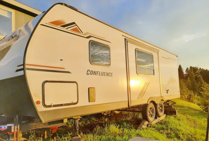 A white 'CONFLUENCE' travel trailer (RV) with mountain decals, parked on grass at sunset. Perfect for an RV rental adventure.. KZ Confluence 2022