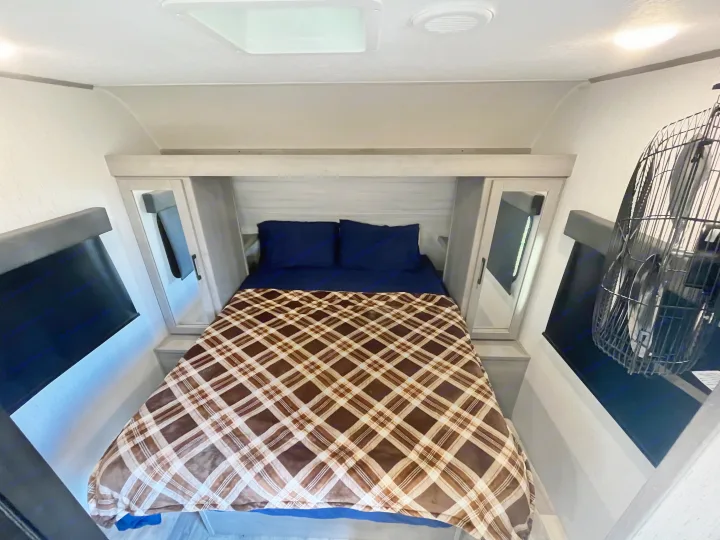 Compact RV rental bedroom with a comfy bed, plaid blanket, bedside mirrors, window shades, and a fan.. Dutchmen Kodiak Bunk House 2019