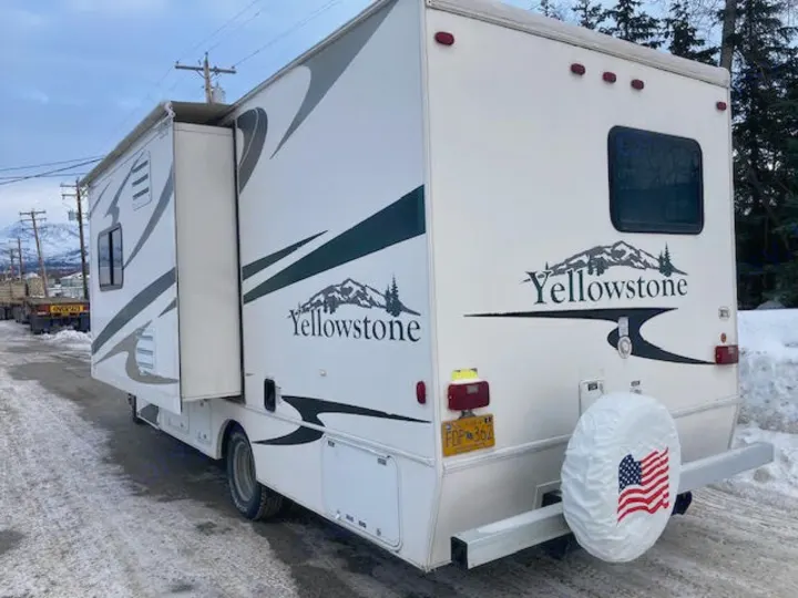 This white Yellowstone RV rental is parked on a snowy road, with its slide-out extended and an American flag on the spare tire cover.. Yellowstone Conquest M6254 2008