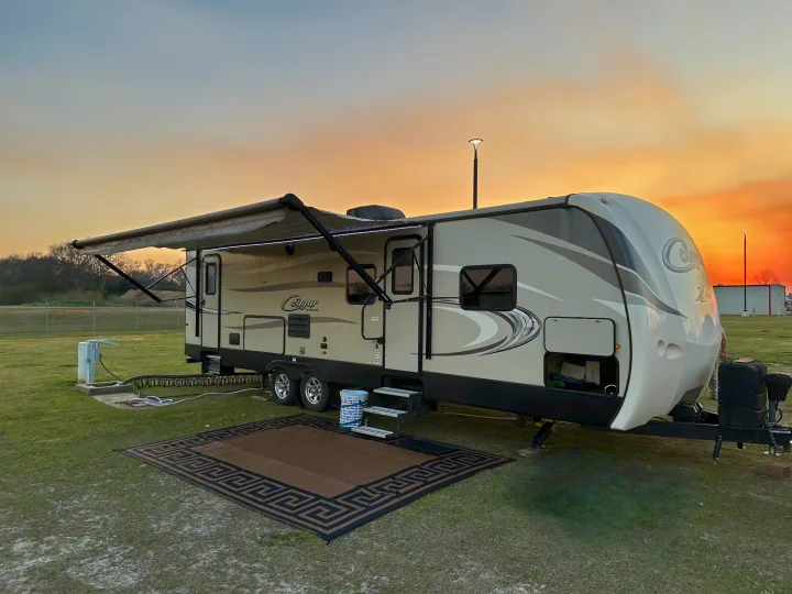 Cozy RV rental campsite at sunset. A light-colored travel trailer with awning extended, outdoor rug, and utility hookups on grass.. Keystone RV Cougar 2019