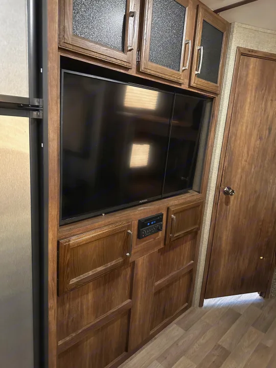 Modern RV rental interior features an entertainment system with TV, stereo, and wood-finish cabinetry beside a refrigerator.. Keystone RV Cougar 2019