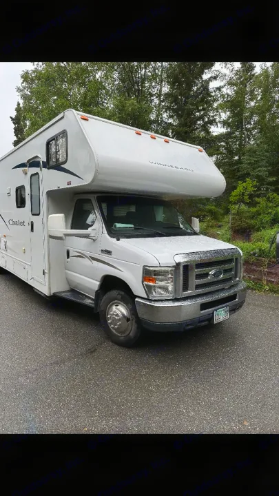A white Winnebago Chalet RV rental is parked on a wet asphalt surface with green trees behind it.. Winnebago Chalet 2010