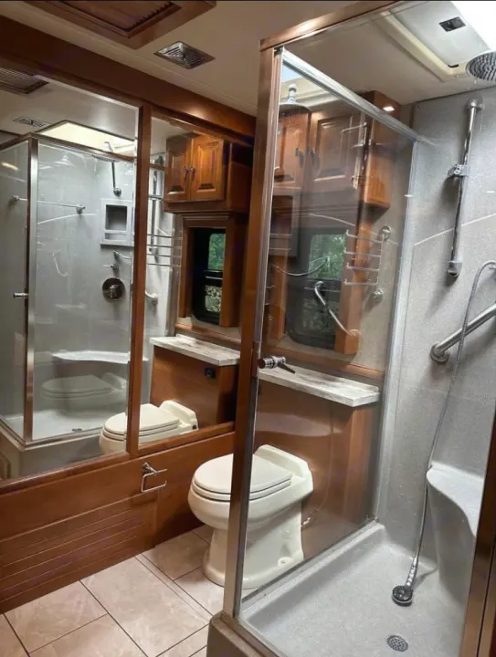 RV rental bathroom with light gray shower stall (right), white toilet, and wooden walls/cabinetry with mirrors.. Tiffin Motorhomes Phaeton 2019