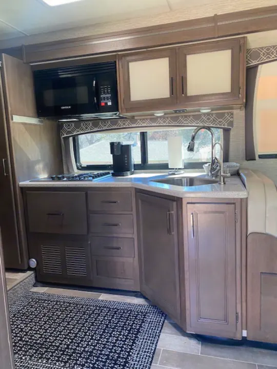 Interior view of an RV rental kitchen showing a black microwave, gas stovetop, stainless steel sink, and wood cabinets.. Thor Chateau 2021