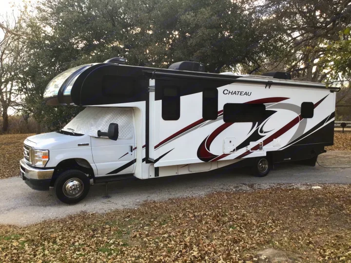 A white and black Chateau Class C RV rental is parked on a gravel lot with trees and autumn leaves, ready for camping.. Thor Chateau 2021