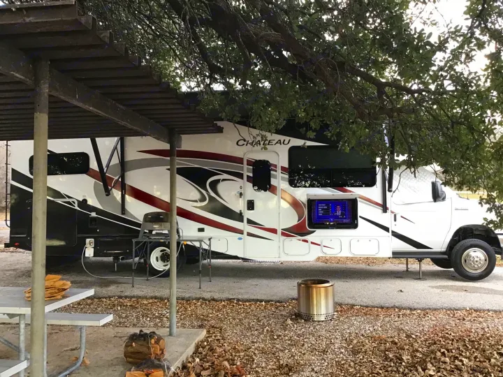 Relaxing at a scenic rv rental campsite with an outdoor TV, grill, and fire pit under the shade of a pavilion.. Thor Chateau 2021