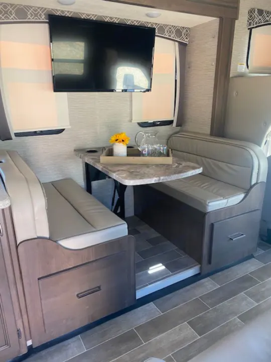 Comfortable RV rental dinette area with built-in benches, a dining table, wall-mounted TV, and wood-look tile flooring.. Thor Chateau 2021