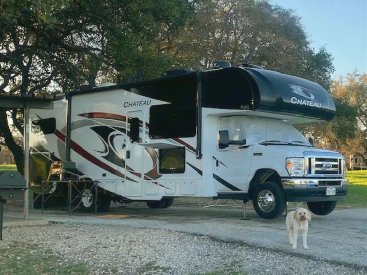 A white Chateau RV rental parked at a campsite, with its awning out, an outdoor cooking setup, and a fluffy dog.. Thor Chateau 2021