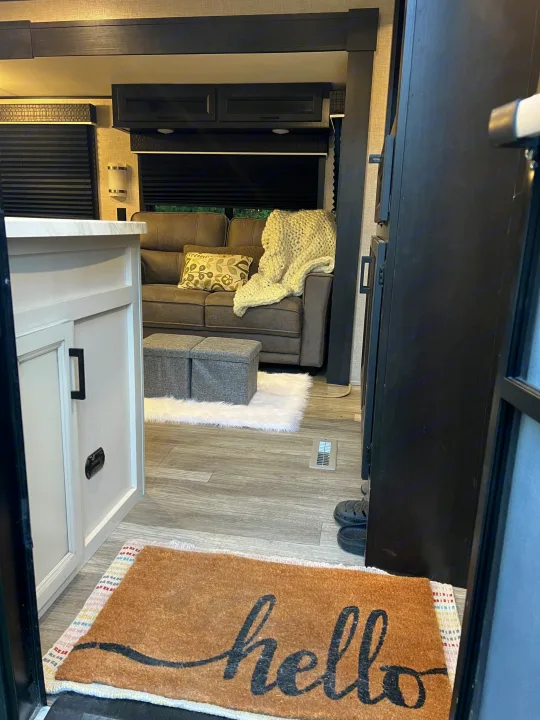 Cozy RV rental living space: a sofa with a chunky knit blanket, ottomans, and a "hello" mat on wood-look floors.. Jayco Jay Flight 2022